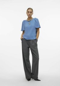 Blusa azzurro chiaro con texture, maniche a palloncino corte, scollatura a chiave, abbinata a pantaloni grigi a vita alta con pieghe e décolleté neri a punta.