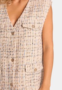 Beige strukturierte Weste mit V-Ausschnitt, die zwei Brusttaschen mit goldenen Knöpfen und blau-braunen gewebten Akzenten aufweist.