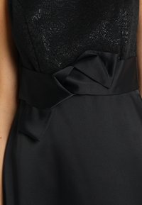 Czarna sukienka z koronkowym, fakturowanym gorsetem, satynowym paskiem w talii z wiązaniem oraz gładkim, zwiewnym materiałem poniżej talii.