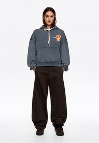 Niebieska bluza z kapturem z przednią kieszenią i graficznym nadrukiem psa, zestawiona z luźnymi czarnymi spodniami i ciemnymi butami. Miękka, teksturowana tkanina.