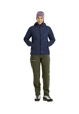 Frau steht und trägt eine marineblaue Kapuzenjacke, olivgrüne Hosen mit Reißverschlusstaschen, Wanderschuhe und ein violettes Stirnband, Hände in den Jackentaschen.