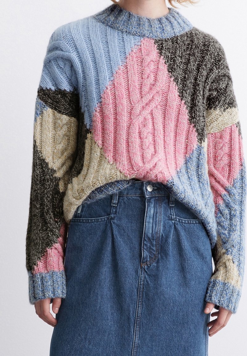 Stickad tröja med en lapptäckesdesign i blå, rosa, brun och beige sektioner; ribbat mönster; hög krage; bärs över en jeanskjol.