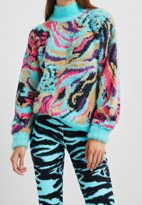 Multifarvet fuzzy sweater med høj hals, med pink, blå og sorte abstrakte mønstre; kombineret med sorte og blå stribede leggings.