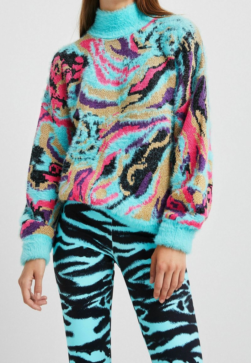 Multifarvet fuzzy sweater med høj hals, med pink, blå og sorte abstrakte mønstre; kombineret med sorte og blå stribede leggings.
