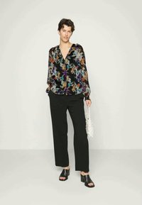 Blusa floral preta com padrões coloridos, frente com botões e mangas longas; combinada com calças pretas e sandálias de salto. Bolsa branca na mão.