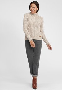 Beige gebreide coltrui met een textuurpatroon, gecombineerd met grijze jeans en bruine enkellaarsjes. Heeft een logo patch aan de zoom.