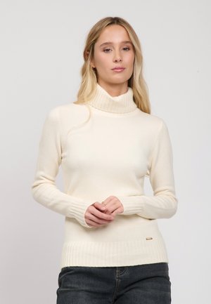 Frau mit langen blonden Haaren, die einen cremefarbenen Rollkragenpullover und dunkle Jeans trägt und vor einem schlichten hellen Hintergrund steht.