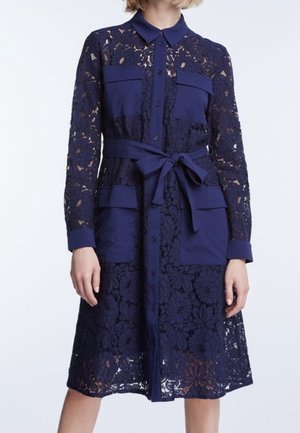 Marineblauwe knielange kanten jurk met lange mouwen, knoopsluiting aan de voorkant, borst- en heupzakken, en een ceintuur die in een strik is gebonden.