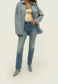 Vrouw in een lichtblauwe denimjas en jeans met een bleekgele top en bijpassende denim haklaarzen, staand met één hand in haar zak.