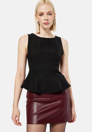 Vaalea nainen, jolla on musta hihaton peplum-toppi ja burgundinsävyinen nahkainen minihame, seisoo yksinkertaisen vaalean taustan edessä.