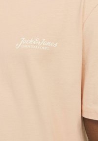 Perzikkleurig katoen T-shirt met korte mouwen, met witte logo tekst "Jack & Jones ESSENTIALS DEPT." op de linkerkant van de borst. Glad van textuur.