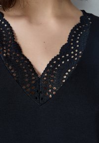 Top nero con scollatura a V profonda rifinita in pizzo, adornato con ritagli circolari lungo il collo per un tocco di texture e dettaglio.