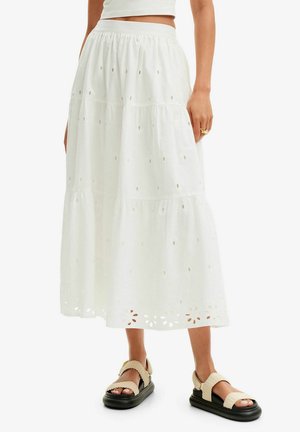 Hvid lagdelt midi øjelet nederdel kombineret med beige teksturerede stroppesandaler på en stående person mod en ensfarvet baggrund.