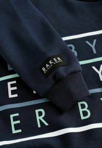 Marineblauer Sweatshirt mit glatter Textur, weißen und hellblauen horizontalen Streifen sowie einem gewebten Etikett am Ärmel mit der Aufschrift "BAKER."