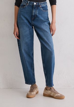 Personne portant un jean bleu taille haute et des baskets beiges, se tenant contre un mur texturé blanc et un sol.