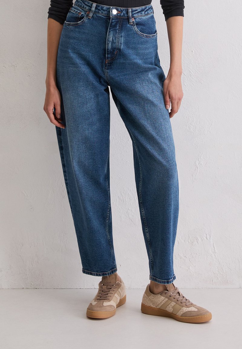 Personne portant un jean bleu taille haute et des baskets beiges, se tenant contre un mur texturé blanc et un sol.