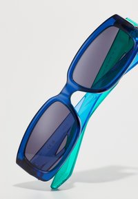 Blå og teal oversized solbriller med mørkt tonede glas og en glat, blank plastikramme. Sekskantede former og teksturerede arme.
