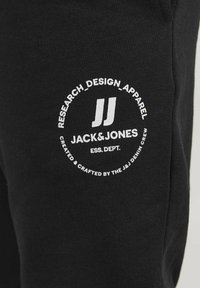 Pantalones deportivos negros con un logo redondo blanco que lleva el texto "RESEARCH_DESIGN_APPAREL" y "JACK&JONES" en el muslo. Tejido suave, diseño casual.