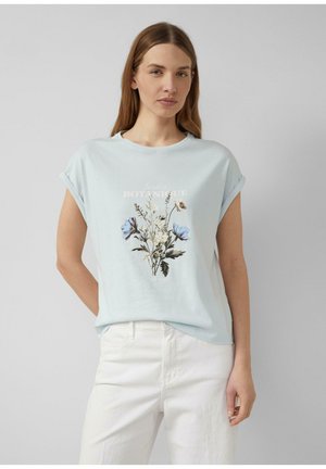 Młoda kobieta o prostych brązowych włosach, ubrana w jasnoniebieską koszulkę z kwiatowym wzorem i białe spodnie, na jednolitym szarym tle.