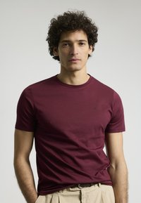 Camiseta corta de manga en color burdeos con cuello redondo, fabricada en tejido suave. Presenta un pequeño logo en el lado izquierdo del pecho.