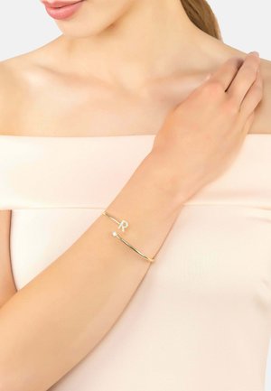 Bracciale rigido in oro con una "R" incisa e un piccolo accento di pietra preziosa trasparente. Finitura liscia e design aperto con un aspetto minimalista.