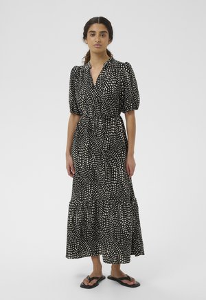 Szőke hajú nő fekete midi ruhában, fehér szabálytalan pöttyök mintázattal és fodros ujjakkal, fekete papucsban, fehér háttér előtt áll.