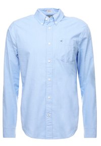 Světle modrá dlouhá košile s knoflíky a s našitým límcem, obsahující kapsu na prsou s malým vyšitým logem.