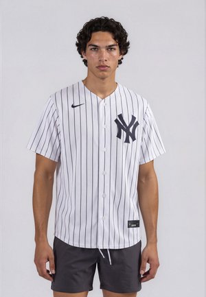 Joven con el pelo rizado que lleva una camiseta de béisbol blanca de los New York Yankees con rayas finas y pantalones cortos negros, de pie frente a un fondo liso.