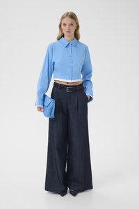 Γυναίκα με κοντό μπλε πουκάμισο με κουμπιά, παντελόνι τζιν σε σκούρο χρώμα με φαρδιά πόδια, μυτερές γόβες και κρατάει μια μπλε τσάντα φλου.