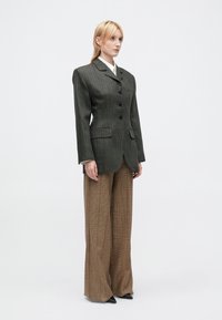 Vrouw die staat en een donkergrijze pinstripe blazer met gepaarde schouders draagt, bruine wijde, geblokte broeken en zwarte puntige schoenen.