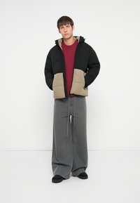 Puffer jacket featuring black upper section and beige lower section, with a hood and pockets. Paired with a maroon shirt and wide grey pants.