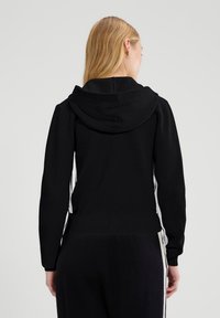 Czarna bluza z kapturem widziana z tyłu, z ribbowanym dołem, długimi rękawami oraz białymi paskami po bokach dla kontrastu. Gładka tekstura materiału.