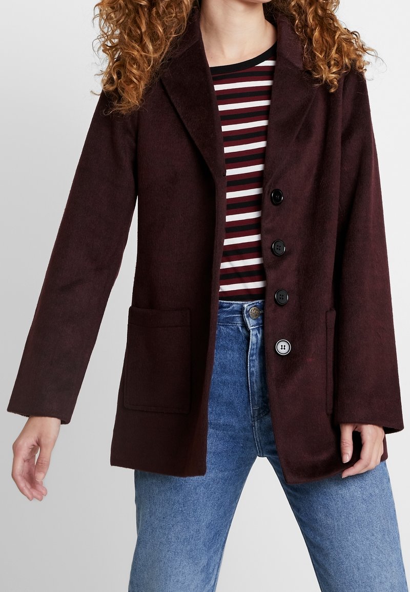 Manteau en mélange de laine bordeaux avec un col cranté, quatre boutons noirs et deux poches avant, associé à une chemise rayée et un jean bleu.