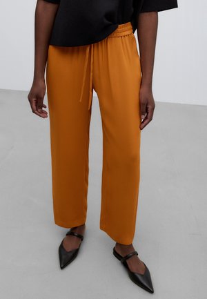 Osoba w musztardowych spodniach o szerokich nogawkach z elastycznym paskiem i czarnych płaskich butach z ostrym noskiem stoi na szarej podłodze.