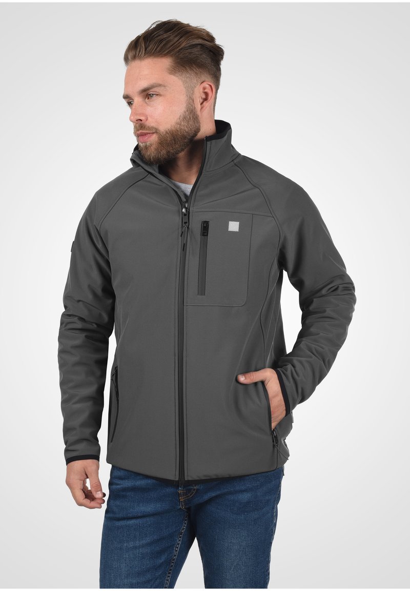 Grijze softshell jas met een ritsborstzak, zijvakken en een hoge kraag. Bevat contrasterende naden en een gladde textuur.