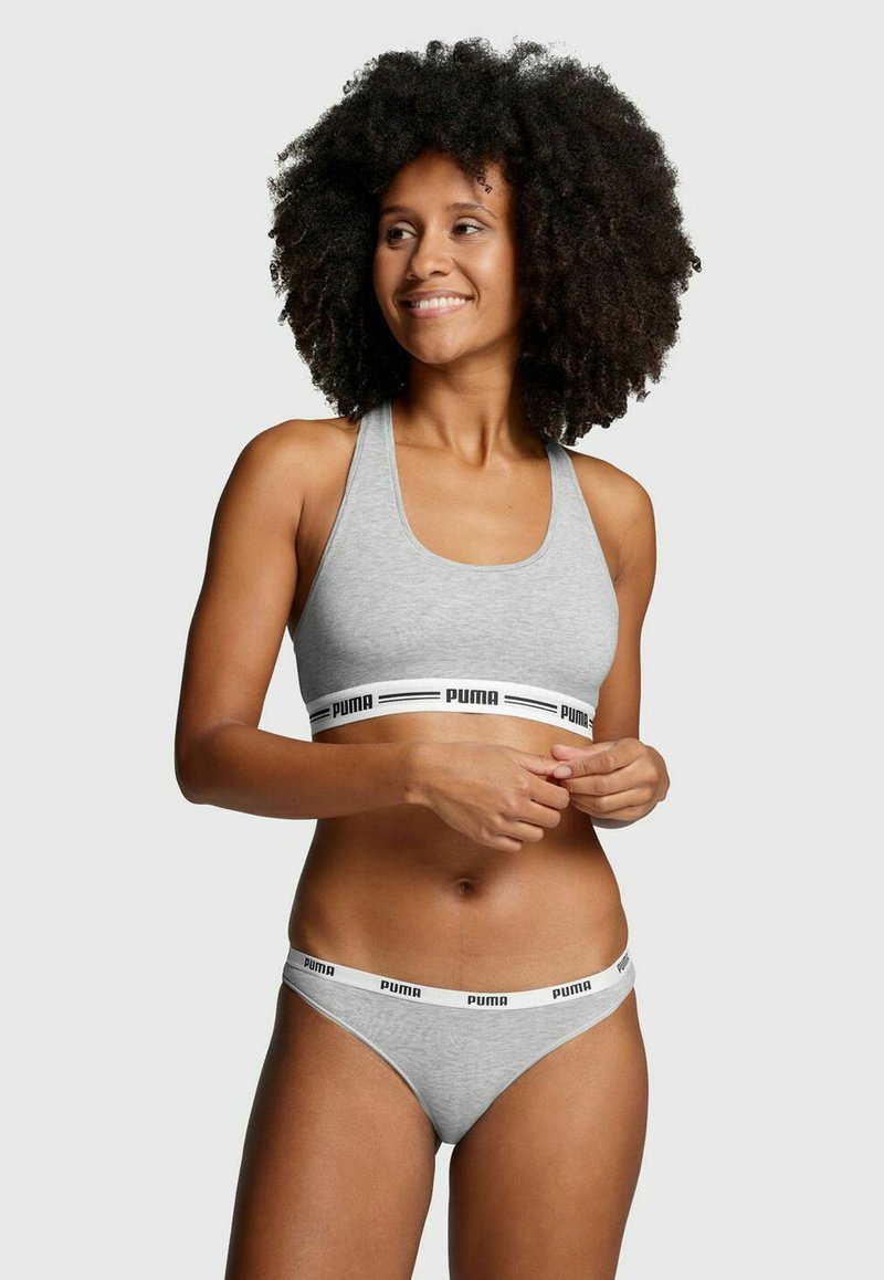Grauer Sport-BH mit einem breiten schwarzen und weißen "PUMA" Logo-Band am unteren Rand; kombiniert mit grauer String-Unterwäsche, die ein ähnliches Banddesign aufweist.