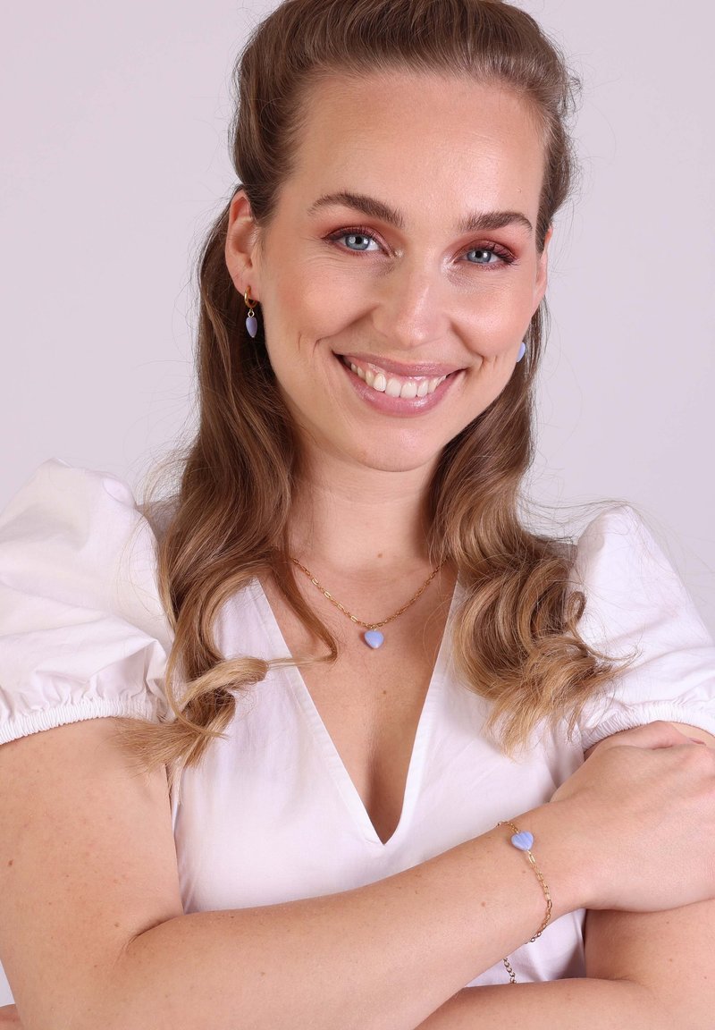Des bijoux en or présentent un pendentif en forme de cœur en pierre bleue sur une chaîne, des boucles d'oreilles assorties, et une blouse à manches bouffantes.