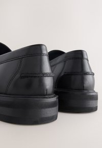 Chaussures en cuir noir présentant un design arrondi et lisse avec des coutures visibles et une semelle épaisse en caoutchouc noir pour une stabilité accrue.