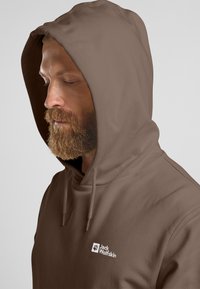 Felpa con cappuccio marrone realizzata in morbido tessuto, con un logo bianco sul davanti. La vista ravvicinata mette in evidenza il cappuccio e il profilo del viso.