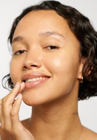 Jeune personne aux cheveux courts et bouclés touchant doucement ses lèvres, souriant légèrement, portant une petite boucle d’oreille et un maquillage minimal, fond uni.
