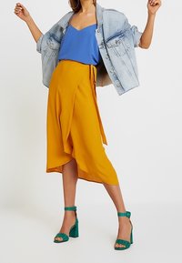 Camisola blu, gonna avvolgente gialla e giacca in denim chiaro. Sandali con tacco verde con cinghie testurizzate e chiusure alla caviglia.