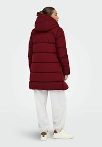 Chaqueta de plumas burdeos con capucha, que presenta un diseño acolchado, mangas largas y hasta las rodillas. Llevada sobre pantalones de chándal grises y zapatillas blancas.
