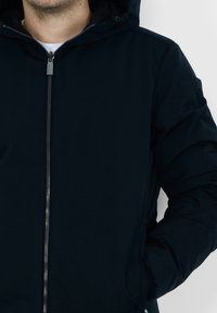Giacca nera con zip e cappuccio, caratterizzata da una superficie liscia e una chiusura con zip frontale, oltre a tasche laterali e un dettaglio del logo sulla manica.