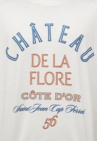 Το κείμενο σε λευκό ύφασμα γράφει «Château de la Flore Côte d'Or Saint Jean Cap Ferrat 56» με μπλε και καφέ στυλιζαρισμένες γραμματοσειρές.