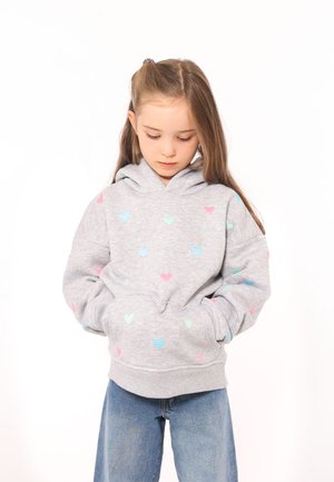 Hellgraue Kapuzenjacke mit einer Fronttasche, die mehrfarbige Herzmuster in Pink und Blau aufweist. Weiches Material und lässiger Schnitt, kombiniert mit blauen Jeans.