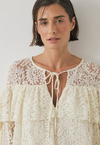 Kermavalkoinen pitsipaita, jossa on röyhelömuotoilu ja avonainen pääntie. Sisältää monimutkaisia kukkakuoseja ja solmimisominaisuuden kaulassa.