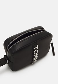Borsa rettangolare nera in pelle con zip argentata, caratterizzata da un audace logo "TOMMY". Texture liscia, design compatto e tracolla regolabile.