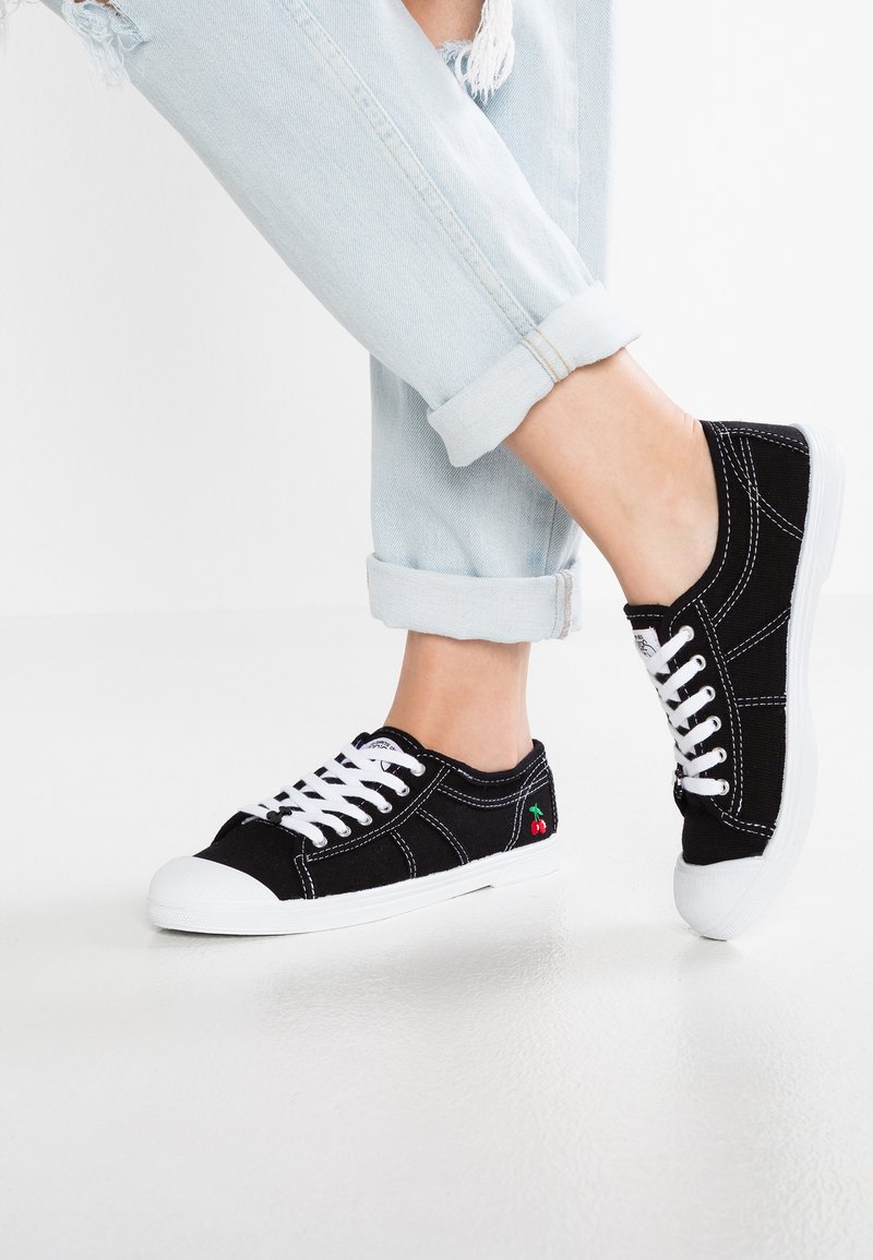 Sneaker in tela nera con lacci e suola bianchi. Presenta cuciture bianche e un piccolo ricamo a forma di ciliegia di lato. Jeans blu risvoltati.