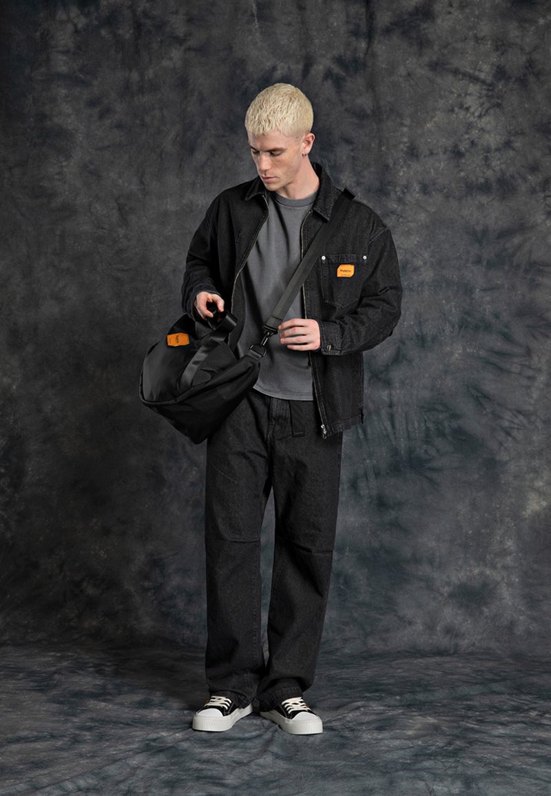 Svart denimjakke med oransje etikett, grå skjorte og løse svarte bukser. Holder en svart skulderveske med justerbar stropp, hvite sneakers.