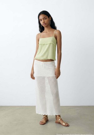 Jeune femme aux longs cheveux noirs porte un haut sans manches vert clair, une jupe maxi en crochet blanche et des sandales dorées à brides, debout sur un sol en béton.